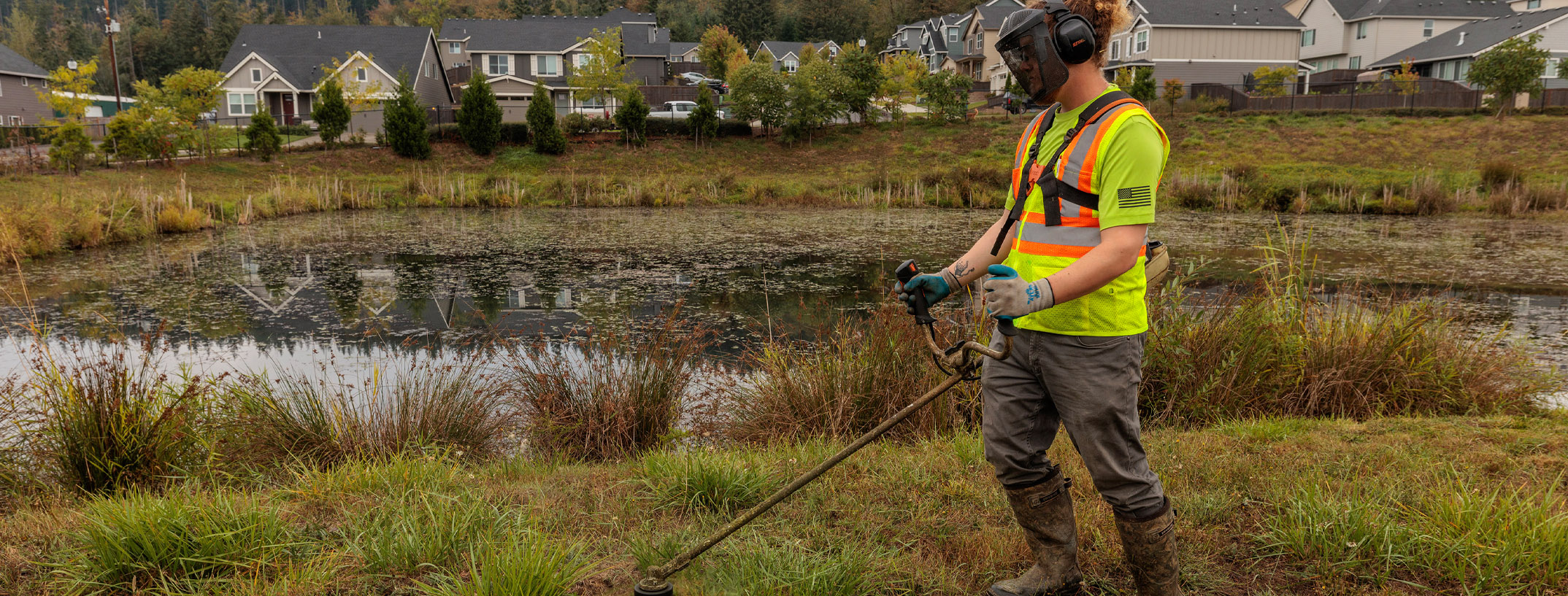 An employee with appropriate safety gear using a weed-eater to remove weeds at the edge of a neighborhood's drainage pond