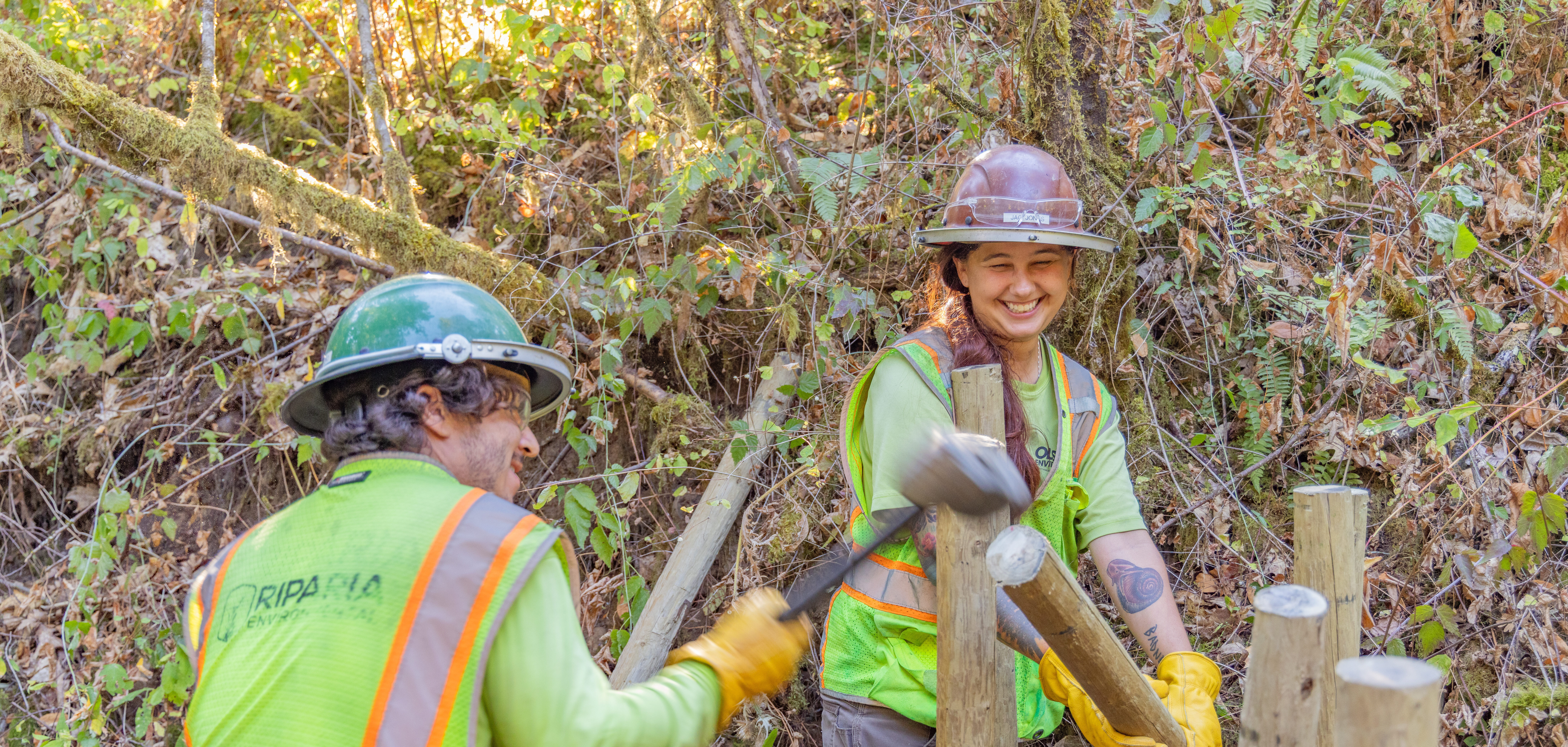 Two trail workers in hard hats and neon vests laugh while hammering wooden posts in a forested hillside.