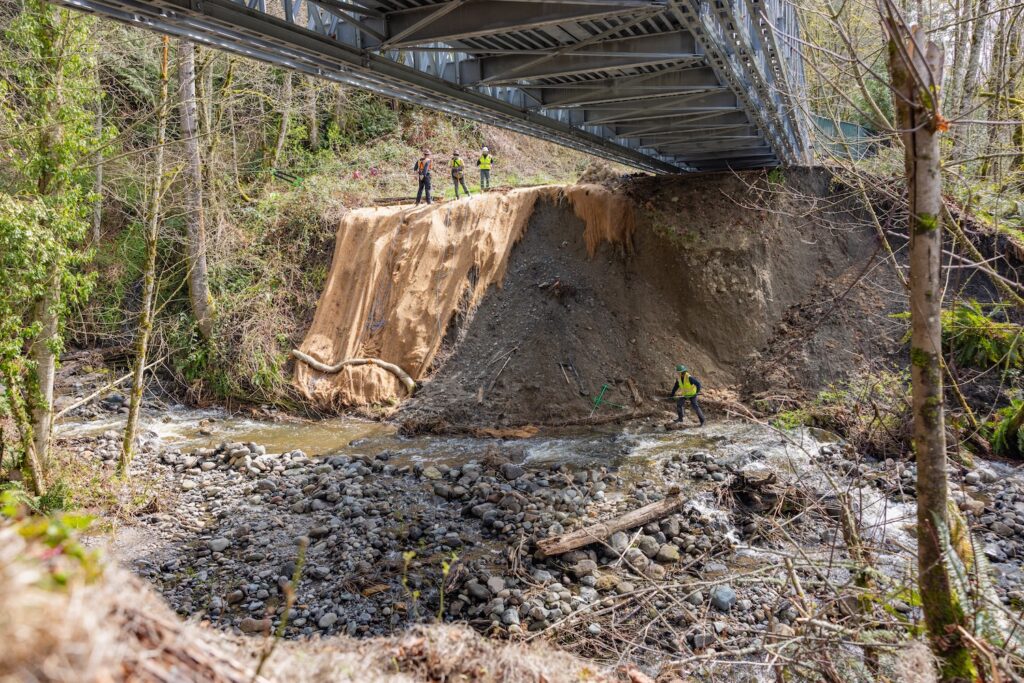 Workers in safety vests inspect a washed-out riverbank under a metal bridge, with one worker crossing the shallow rocky stream.
