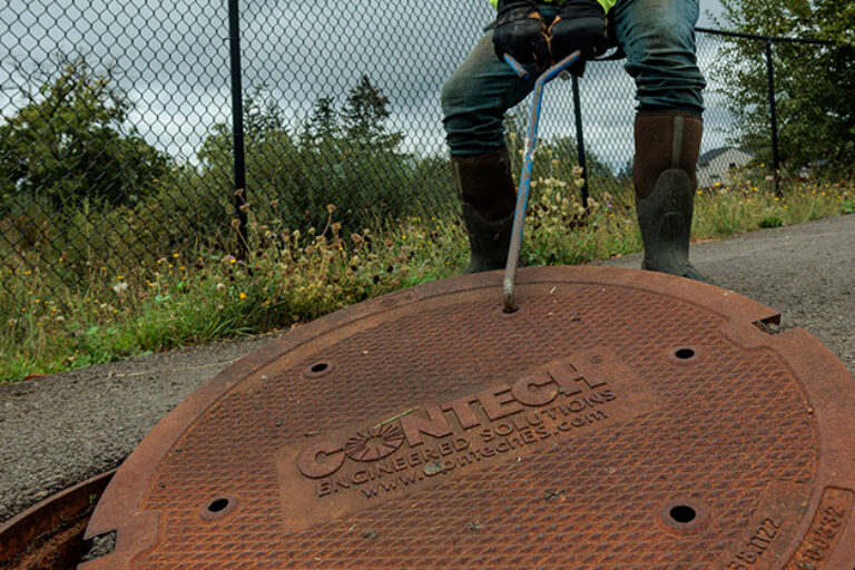 Worker in boots lifting a rusted cast-iron manhole cover labeled "Coaltech" with a long metal hook by a chain-link fence