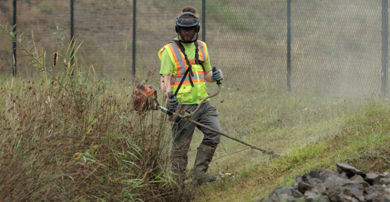 A worker in protective gear using a weedeater to cut weeds and grass along the edge of a natural trail