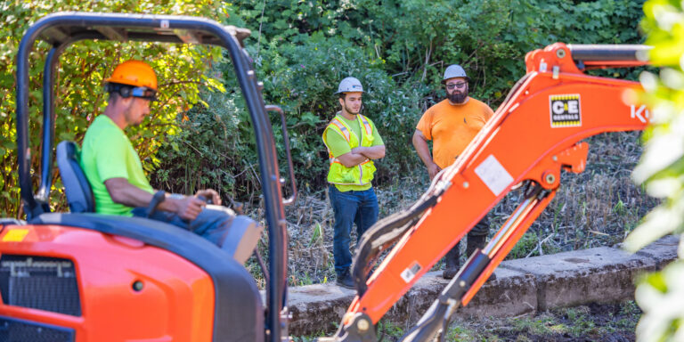 Three construction workers in hard hats and hi-vis clothing standing near an orange excavator in a leafy worksite.