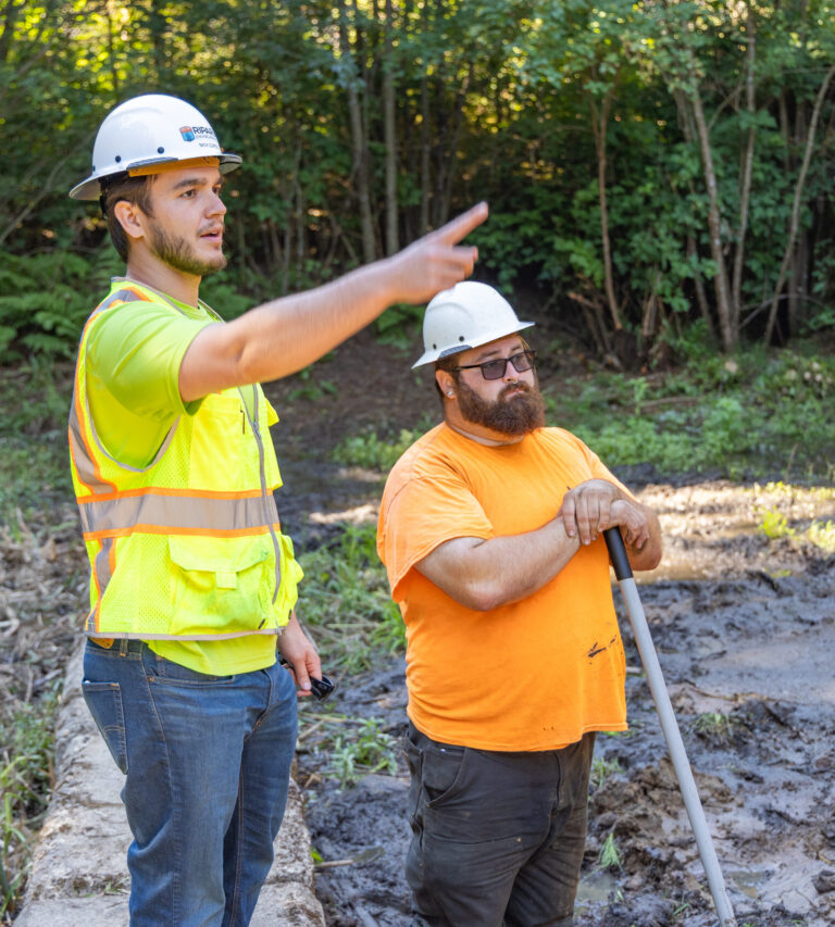 Two construction workers in hard hats and high-visibility shirts stand by a muddy site, one pointing while the other rests on a shovel.
