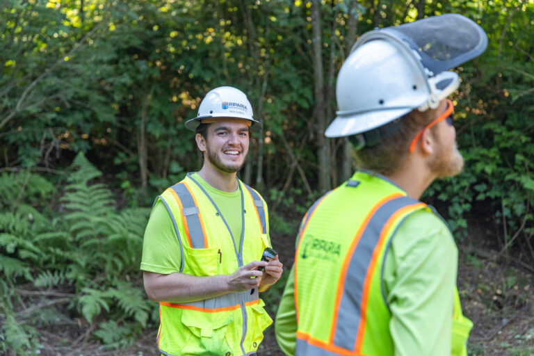Construction workers in high-visibility vests and white hard hats standing in a wooded area, one smiling toward the camera.
