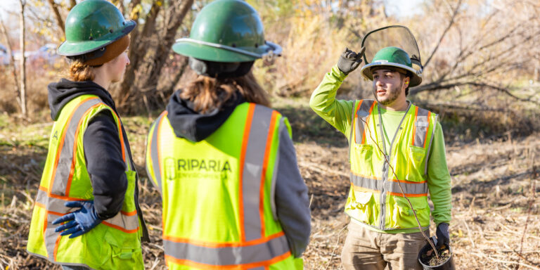 Three workers in green hard hats and high-visibility vests stand outdoors; one man lifts a face shield while holding a small plant pot.