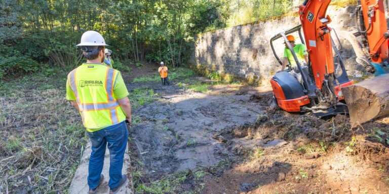 Workers in safety vests and hard hats stand beside a muddy excavation while an orange Kubota mini excavator operates near a stone wall