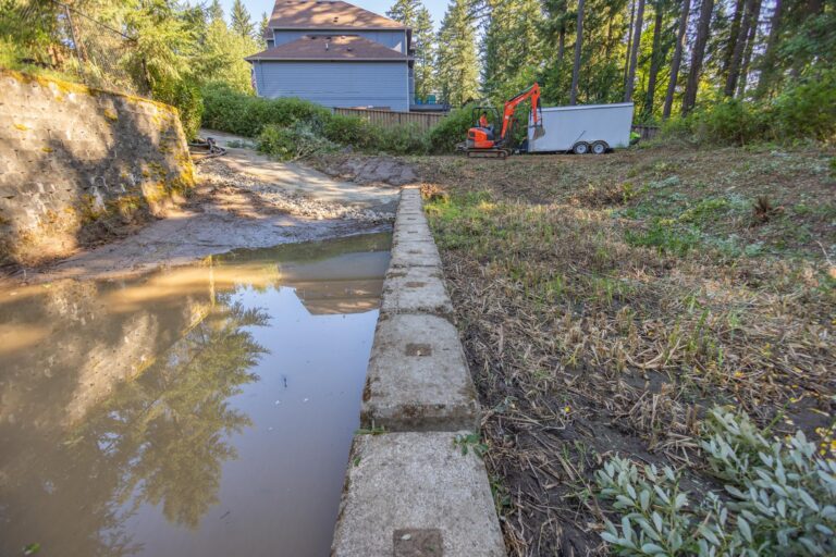 Narrow concrete retaining wall beside a muddy pond with excavator, trailer, cleared vegetation and a blue house in background