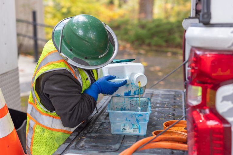 Worker in green hard hat and reflective vest pouring yellow liquid from a white jug into a clear container on a truck bed