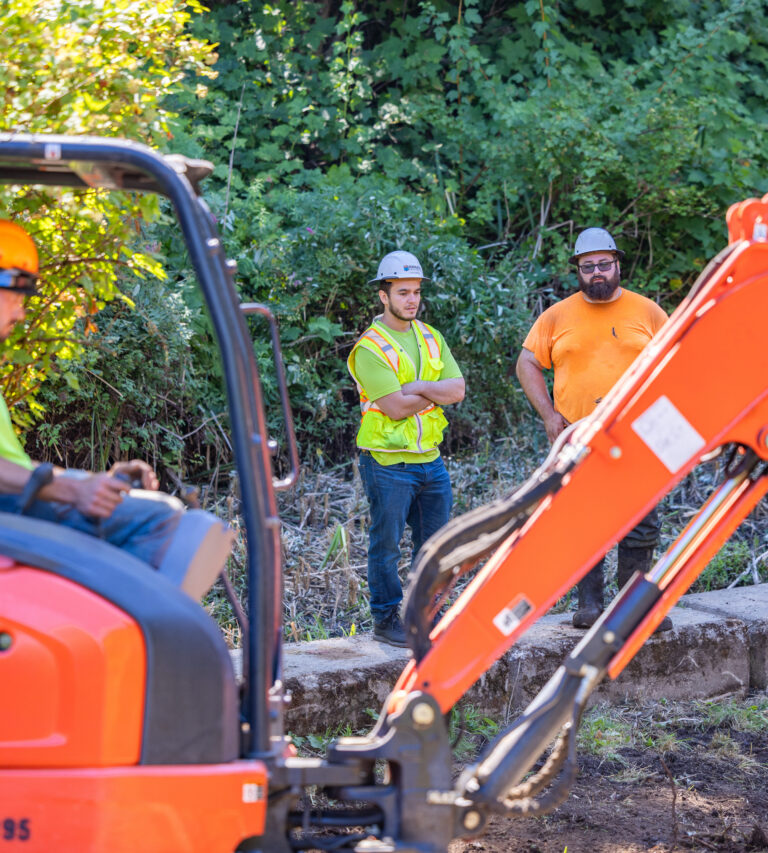 Three construction workers in hard hats and safety vests standing by an orange excavator in a leafy worksite.