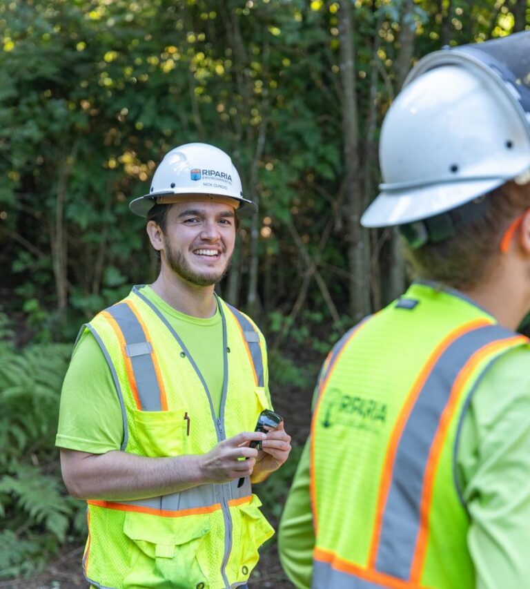 Two construction workers in neon safety vests and white hard hats standing outdoors among trees, one smiling at camera while holding a device.