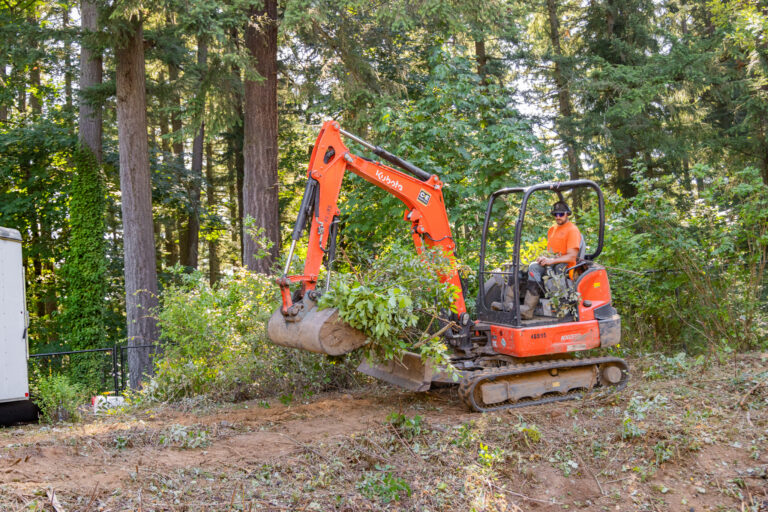 Worker in orange shirt operating a small orange excavator clearing brush in a wooded area
