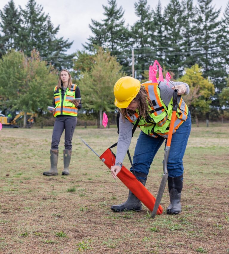 Two utility workers in high-visibility vests and hard hats placing pink-flagged stakes in a grassy field