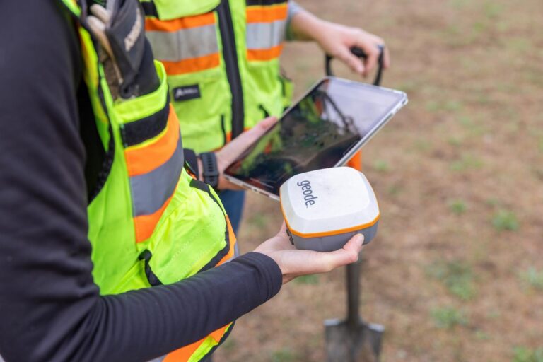 Worker in high-visibility vest holding a Geode GNSS receiver while a colleague views a tablet and a shovel rests nearby