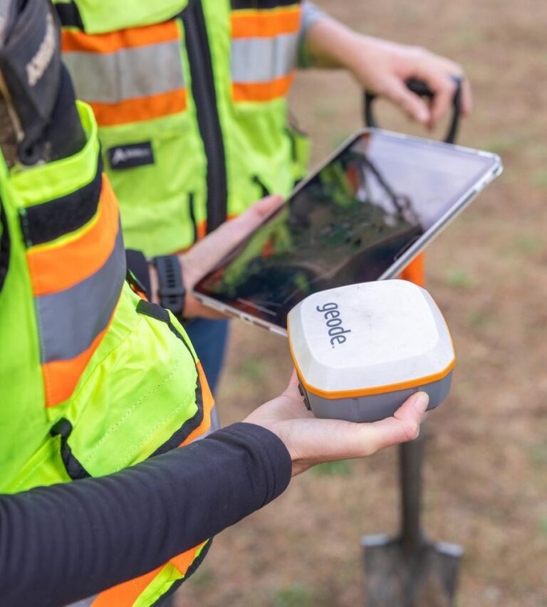 Worker in high-visibility vest holding a Geode GNSS receiver while a colleague views a tablet and a shovel rests nearby