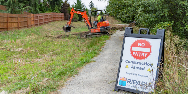 No entry construction sign on a gravel path with an orange mini excavator and operator digging beside a wooden fence and trees