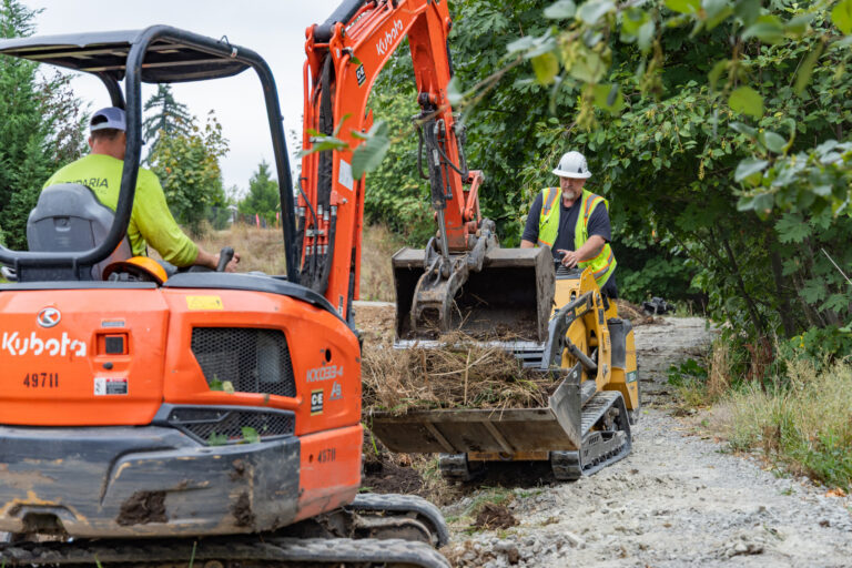 Construction workers in safety vests and helmets operating an orange excavator and a small loader clearing brush on a dirt path