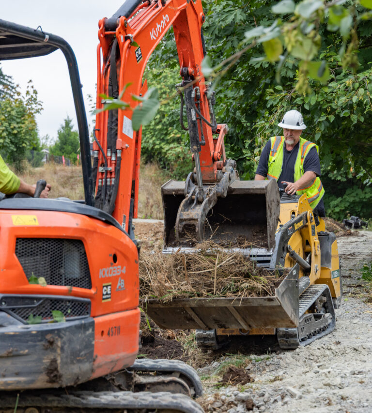 Construction workers in safety vests and helmets operating an orange excavator and a small loader clearing brush on a dirt path