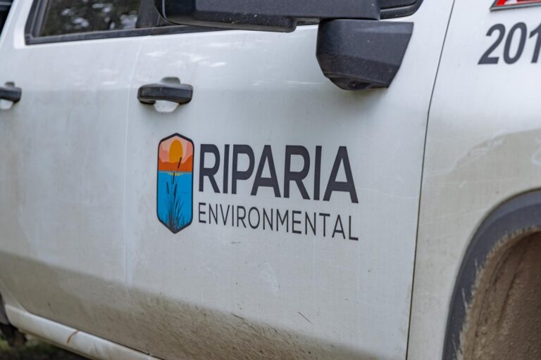 White work truck door with a colorful shield logo and text reading "RIPARIA ENVIRONMENTAL," dirt splashes along the lower panel.
