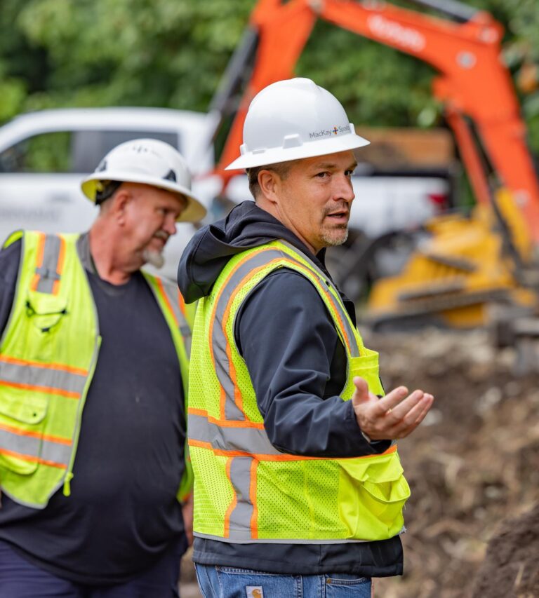 Construction foreman in white hard hat and neon safety vest gesturing forward with coworker and excavator in background