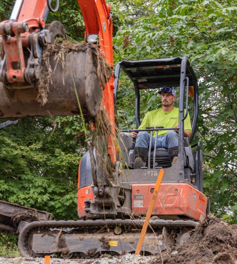 Construction worker in a neon shirt operating an orange excavator, bucket lifting soil and roots at a wooded excavation site.