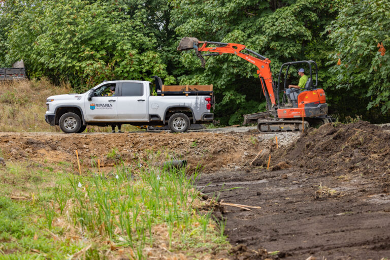 White Riparia Environmental pickup beside an orange mini excavator loading dirt, worker operating machine near wooded background
