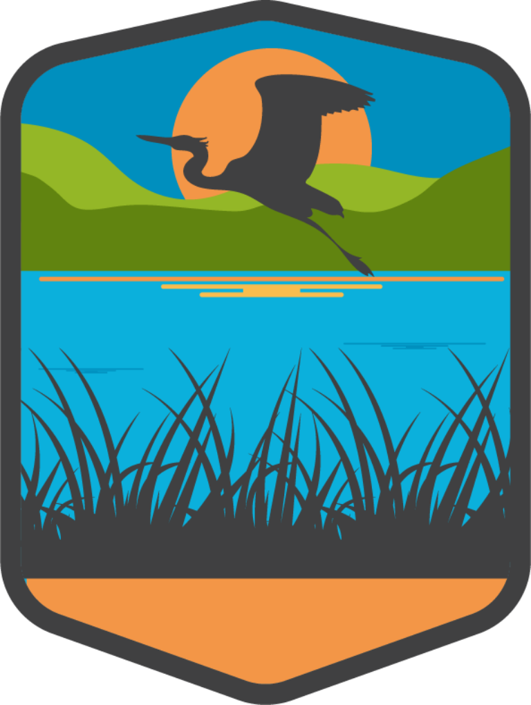 Stylized shield-shaped badge showing a heron silhouette flying over a blue lake with reeds in the foreground, green hills and an orange sun in the background.