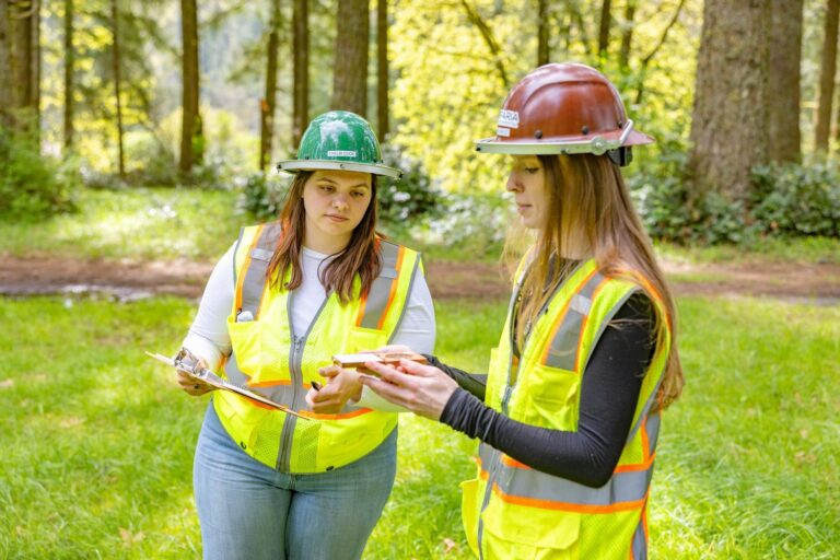 Two women in hard hats and reflective vests standing in a grassy forest clearing, one holding a clipboard while the other shows a small sample.