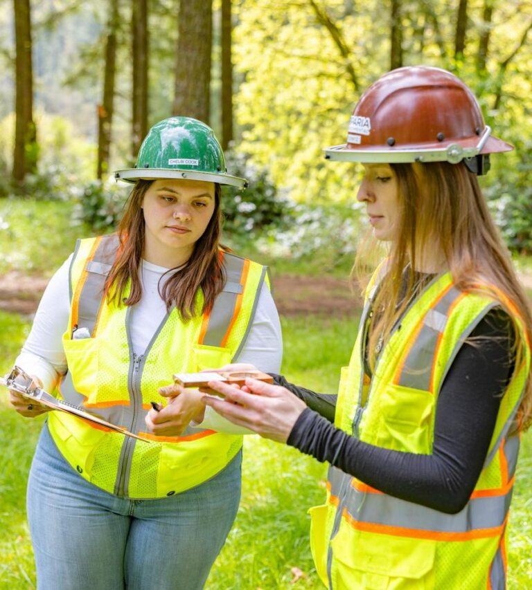 Two women in hard hats and reflective vests standing in a grassy forest clearing, one holding a clipboard while the other shows a small sample.