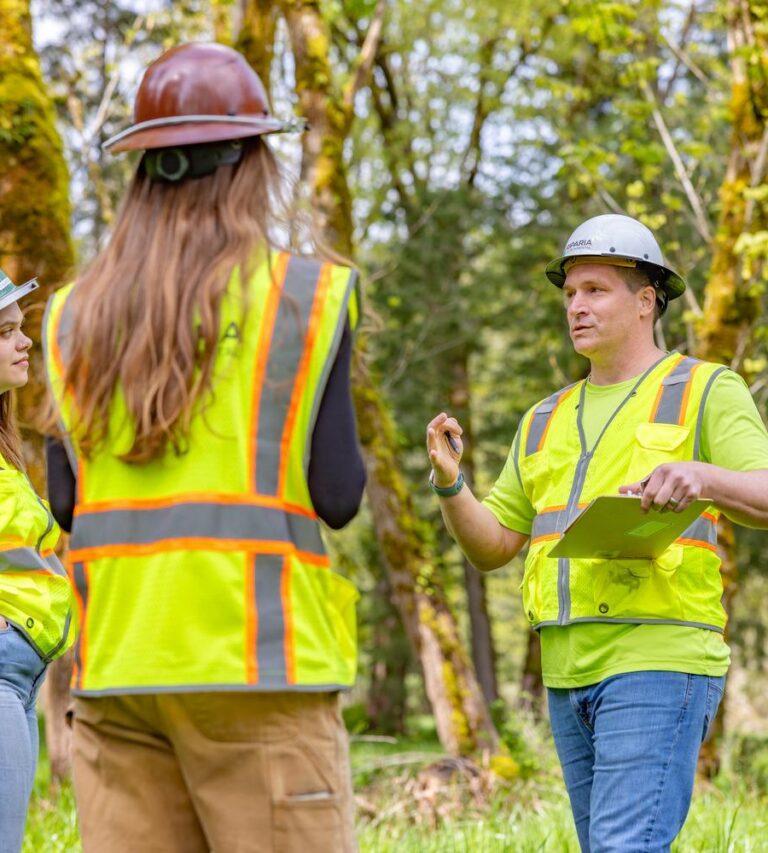 Three construction workers in safety vests and hard hats talking in a wooded outdoor worksite, one holding a clipboard.