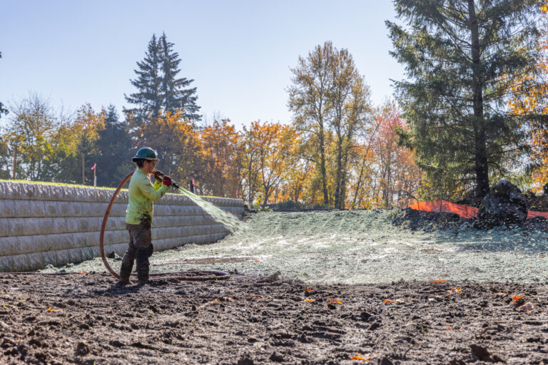 Worker in a hard hat spraying green hydroseed over a muddy slope with autumn trees in the background