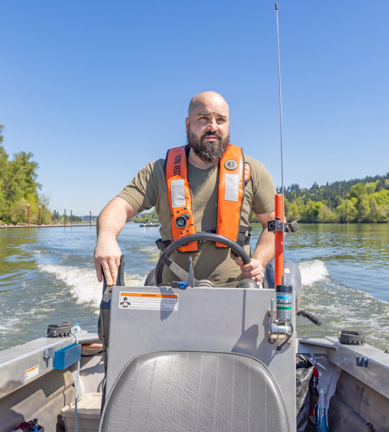 Bearded man wearing an orange life vest driving a small motorboat on a sunny river with tree-lined banks.