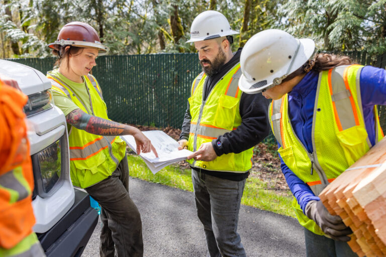Three construction workers in hard hats and high-visibility vests study plans while another worker holds stacked wood.