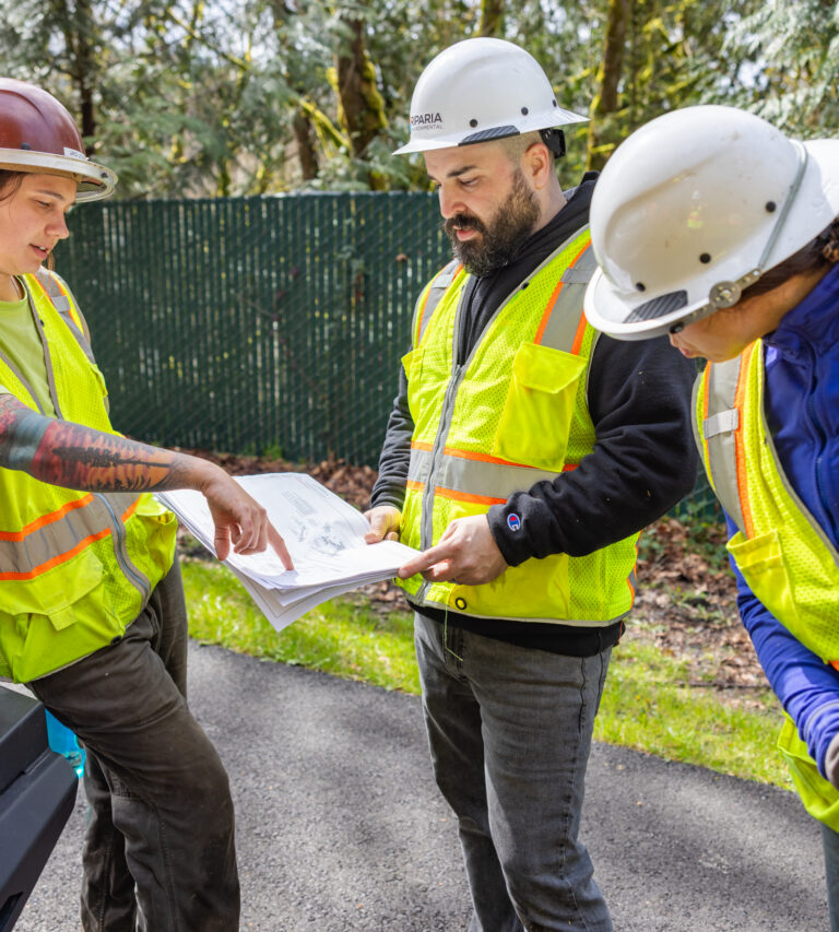 Three construction workers in hard hats and high-visibility vests study plans while another worker holds stacked wood.