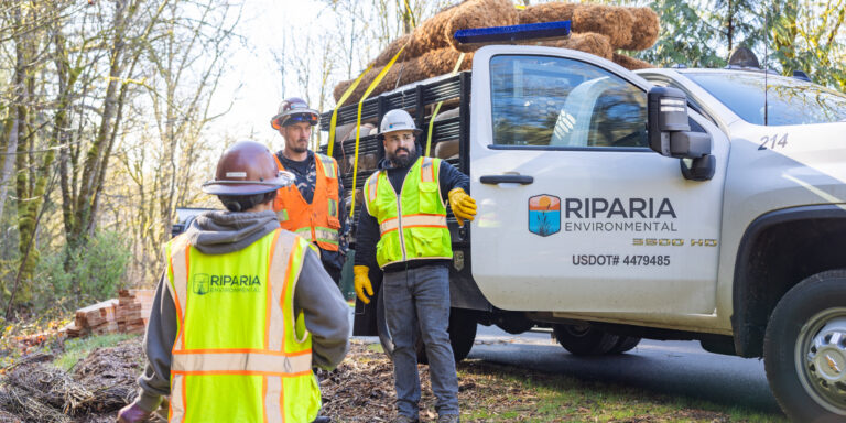 Three workers in high-visibility vests and hard hats standing beside a Riparia Environmental pickup truck loaded with coir rolls