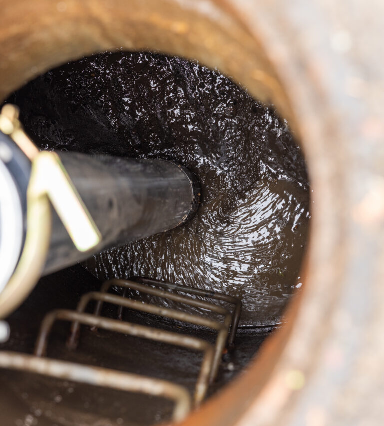 View down into a manhole showing murky wastewater, a vertical pipe, and metal ladder rungs along the wall
