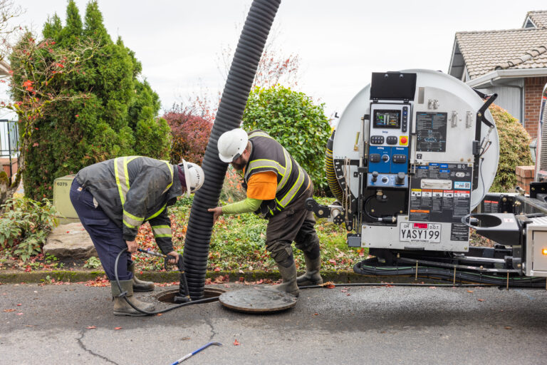 Two utility workers in safety vests and hard hats feeding a large corrugated vacuum hose into an open manhole beside a service truck.