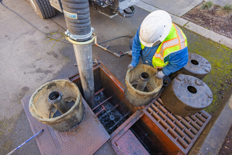 Worker in hard hat and high-vis vest removing debris filters from an open storm drain with suction hose and truck nearby