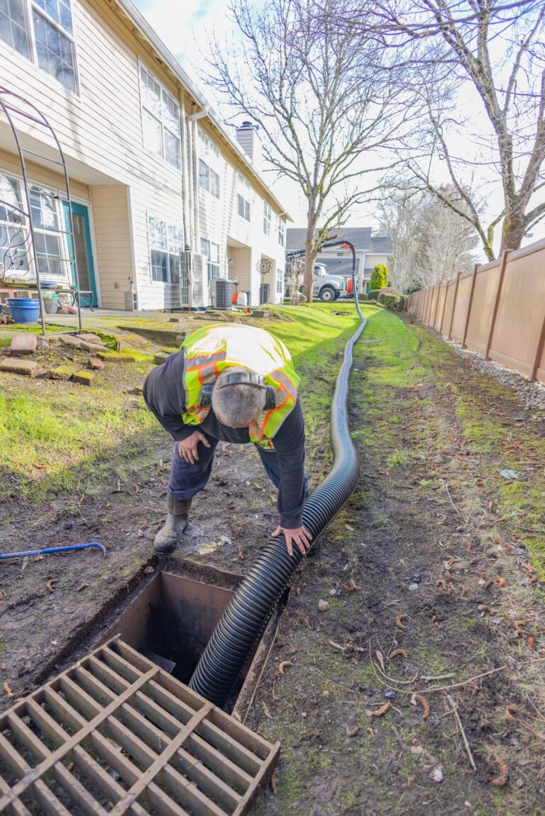 Worker in a safety vest bending to feed a large black corrugated drainage hose into a storm drain beside townhouses