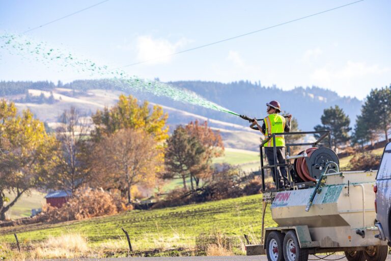 Worker in a high-visibility vest spraying green slurry from a hose mounted on a trailer in a rural landscape