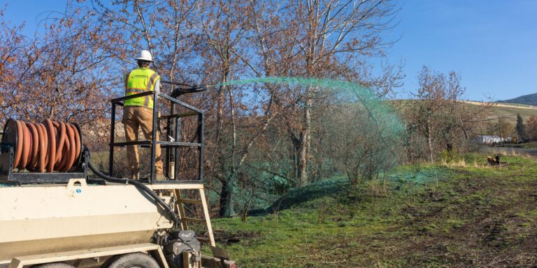 Worker in a hard hat and high-visibility vest spraying green hydroseeding slurry from a tanker onto a grassy hillside with bare trees.