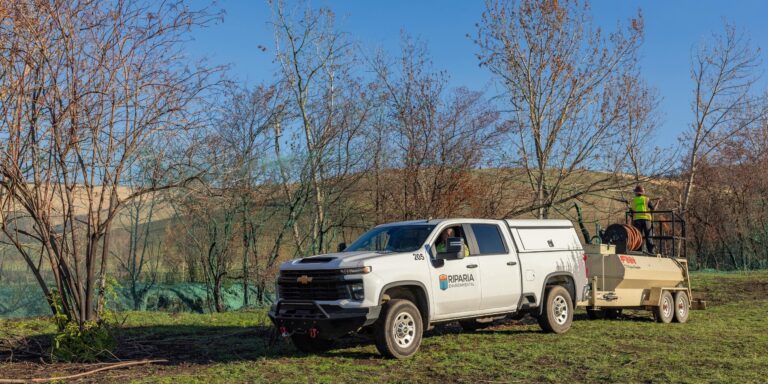 White Riparia Environmental pickup towing equipment while a worker in a high-visibility vest stands on the trailer among leafless trees.