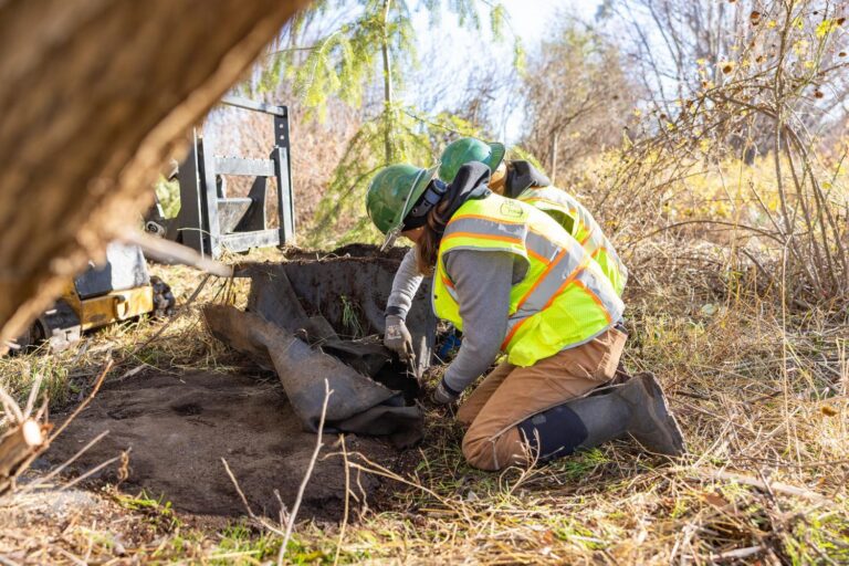 Two workers in green hard hats and reflective vests kneel outdoors, removing turf and fabric from a dug patch of soil.