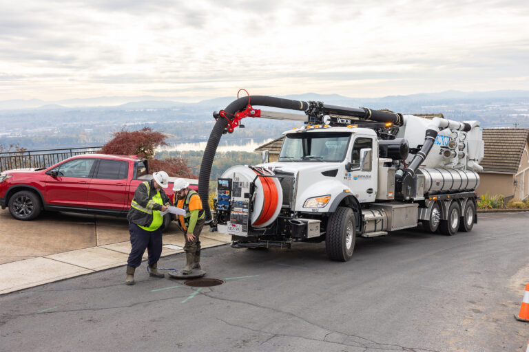 Two workers in safety gear inspecting plans beside an open manhole and a large vacuum truck with extended hose on a hillside road