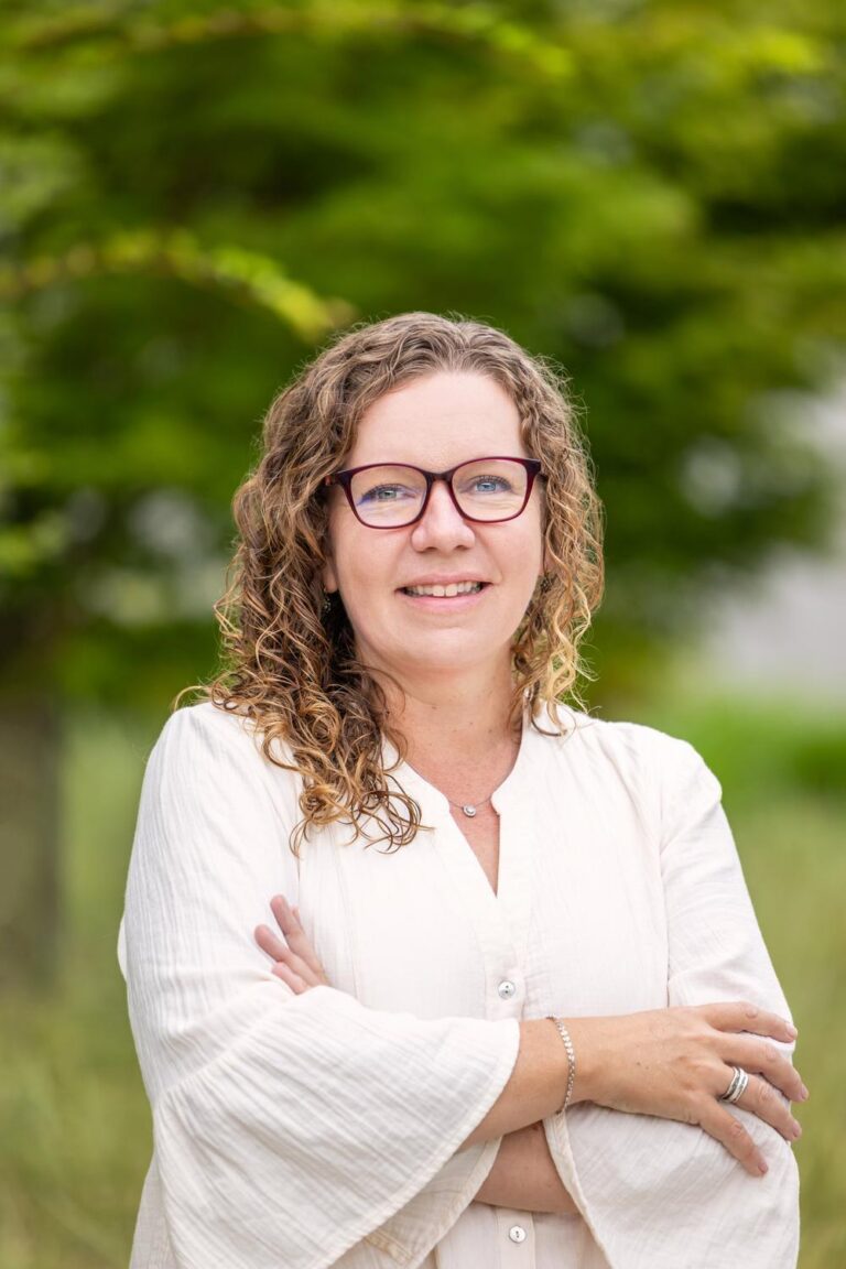 Smiling woman with curly hair and red-framed glasses wearing a light blouse, arms crossed, standing outdoors with blurred green foliage behind her