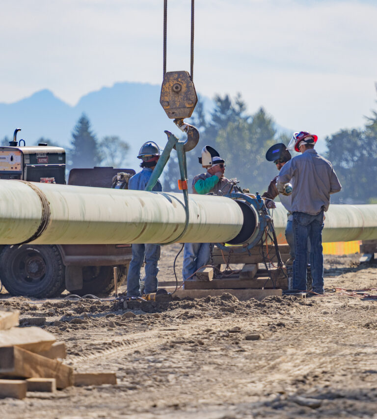 Workers in hard hats and welding masks guide a large green pipeline section suspended by a crane hook at a dusty construction site.