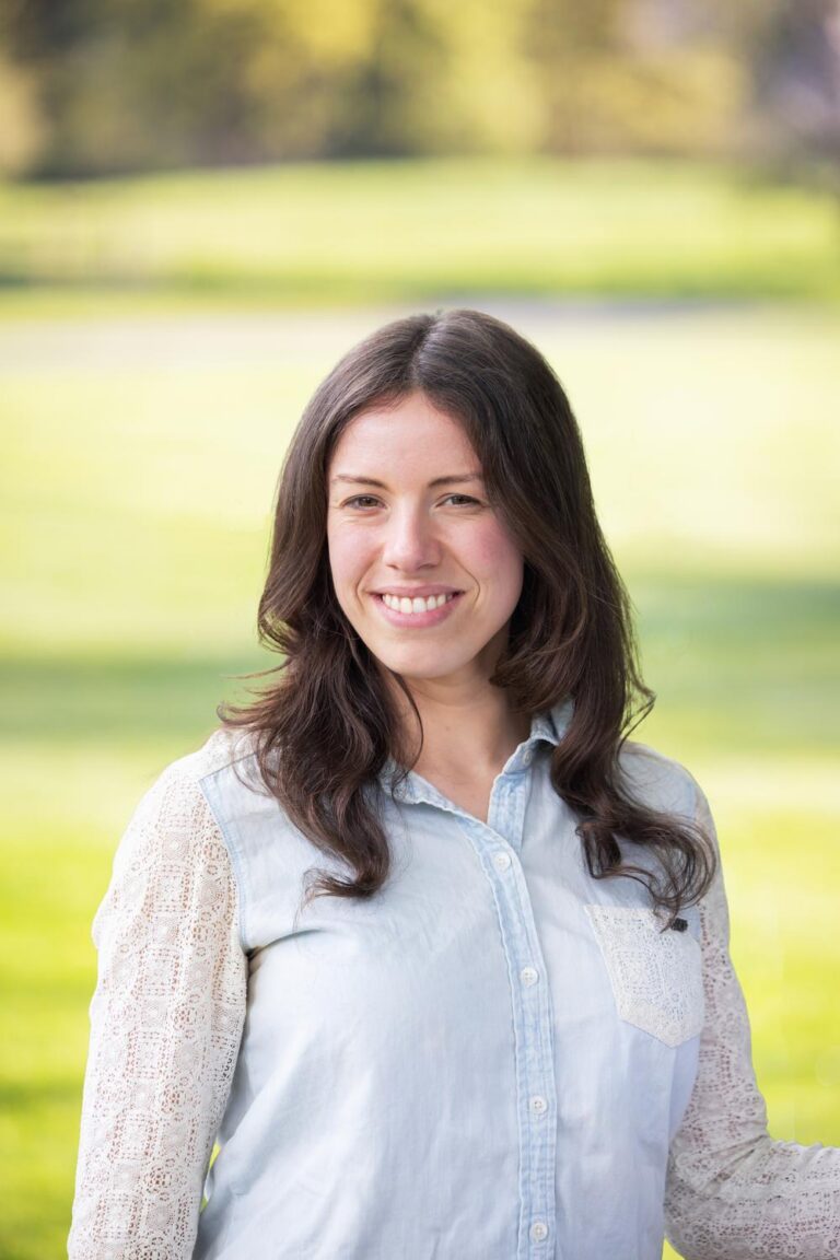 Smiling young woman with long dark hair wearing a light denim shirt with lace sleeves, standing outdoors on a sunny grassy background