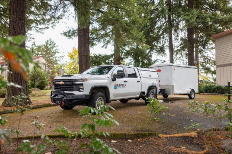 White Riparia Environmental pickup truck parked on a tree-lined driveway hauling a white enclosed trailer.