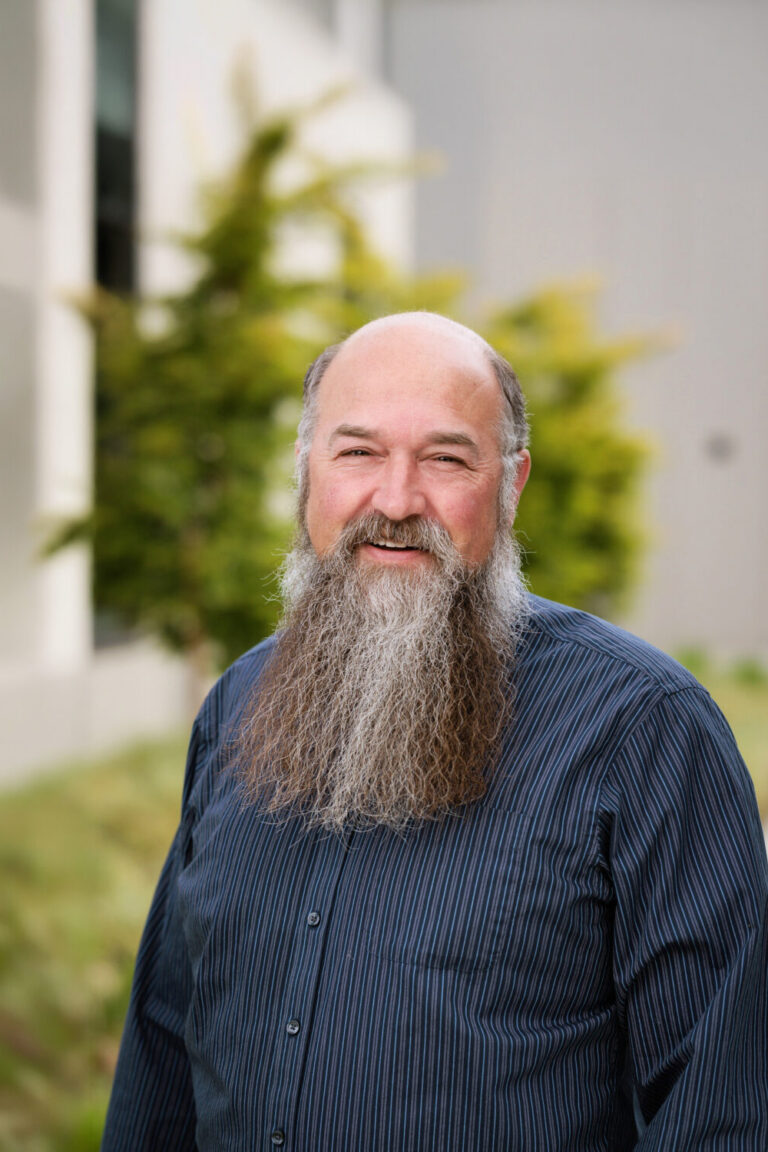 smiling middle-aged man with a long gray beard, balding head, and striped dark button-up shirt standing outdoors with blurred greenery behind him