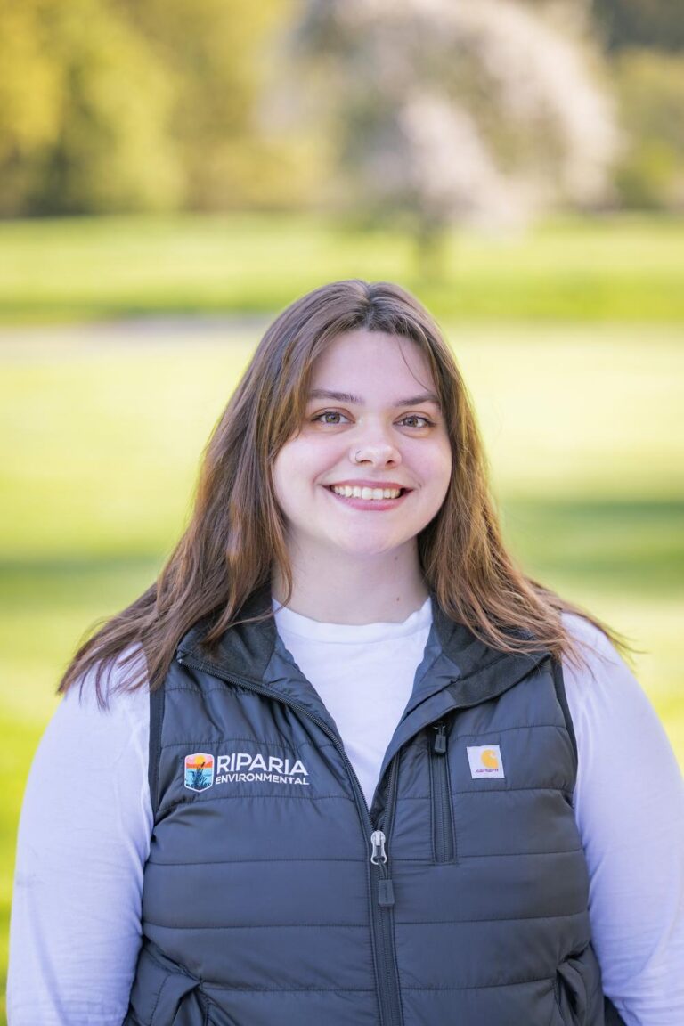 Young woman smiling outdoors wearing a black Riparia Environmental puffer vest over a white shirt with green park background