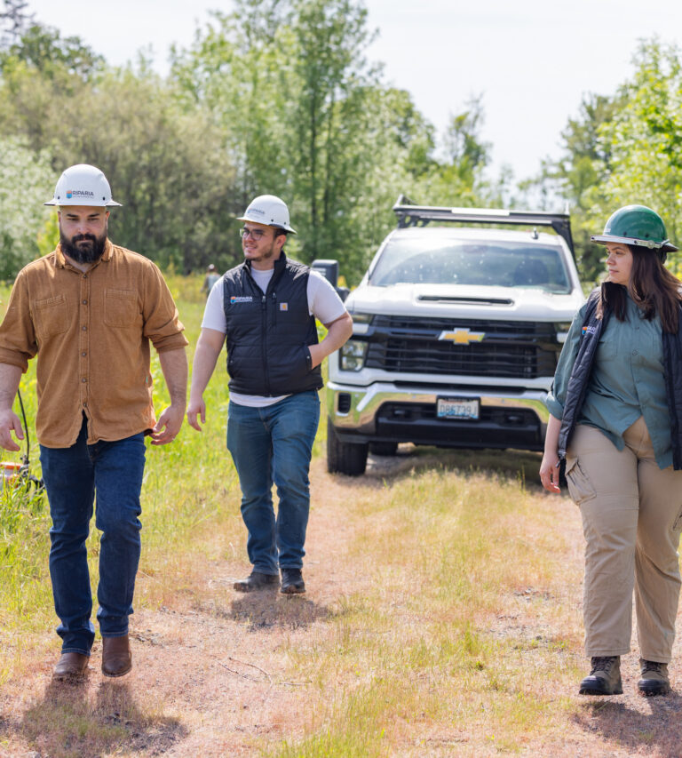 Three field workers wearing hard hats and vests walk along a dirt path in a grassy, tree-lined area with a white pickup truck behind them.